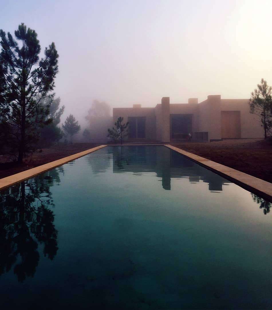Long pool reflecting a sand-colored house in morning mist
