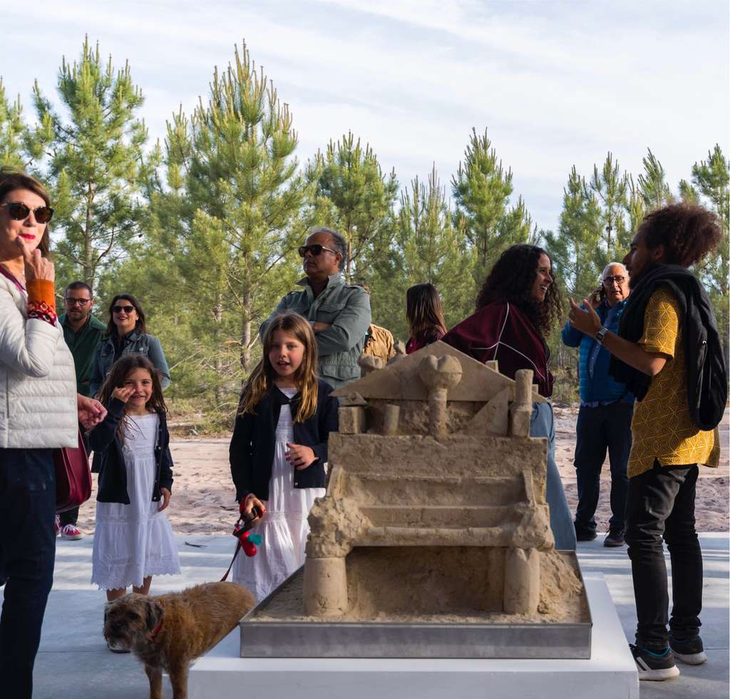 Visitors, children and a dog gather around a sand sculpture of a chair
