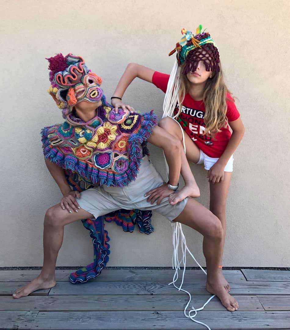 Two people in crocheted masks and costumes posing against a wall