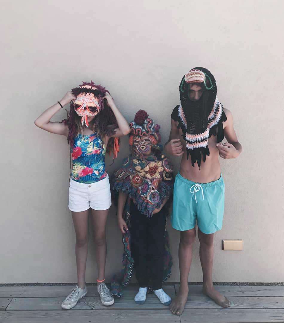 Three people with crocheted masks covering their faces against a wall