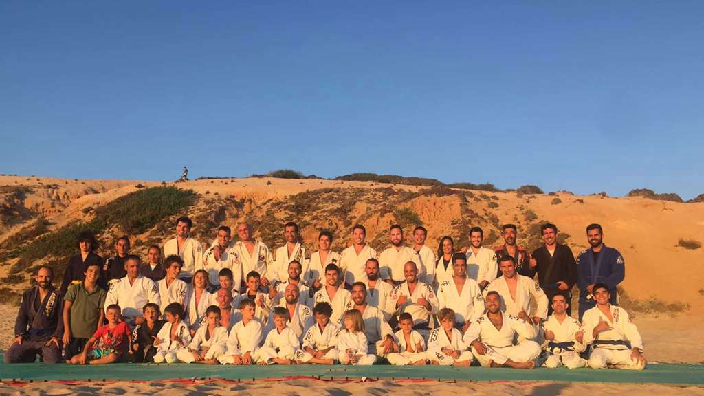 Large group in white gis posing on a beach with orange cliffs behind