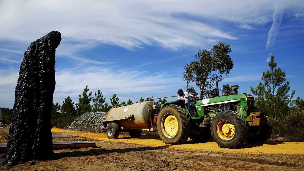 Black rough-textured sculpture beside a green tractor with a tank on a sandy track