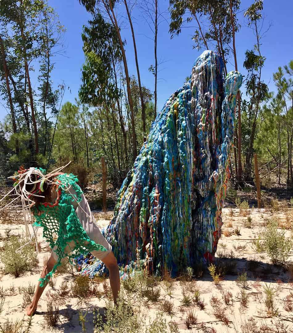 Person in a green crocheted costume leaning beside a tall blue woven sculpture